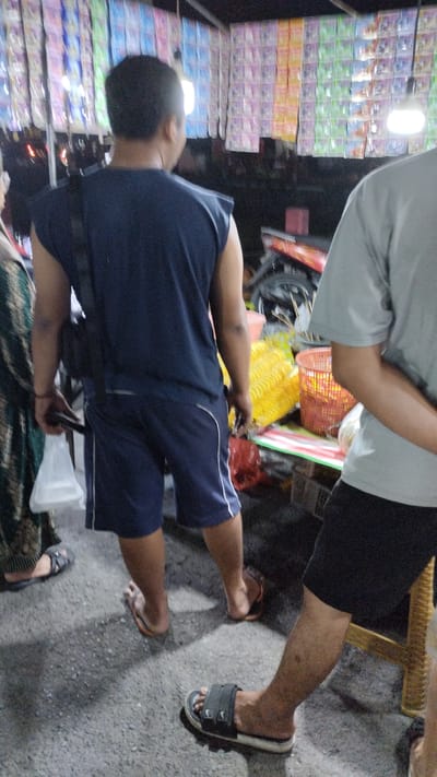 The image captures a nighttime scene at a street-side vendor stall in Sukodono, Indonesia. The stall displays a variety of brightly packaged goods hanging from the ceiling, illuminated by fluorescent lights. In the foreground, a person wearing a dark blue sleeveless shirt and navy shorts walks away from the camera, carrying a white plastic bag in their left hand. Another individual, partially visible from the back, wears a light grey t-shirt and black shorts, standing near a wooden stool. Several other people are present but mostly out of focus. The ground is paved with asphalt. On the vendor's table, there are displays of yellow spiral-shaped snacks and items in a red basket. A motorcycle is parked behind the stall.