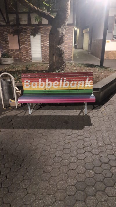 The image shows a rainbow-colored bench with "Babbelbank" written on it in white letters. The bench is located on a hexagonal stone pavement in the city of Bingen am Rhein, Germany. A tree is to the left of the bench, and a trash can is also visible. The background includes a brick building and a lamppost. The scene appears to be outdoors at night.