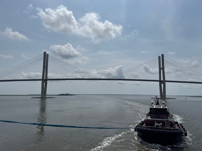 The image depicts a scene on the Brunswick River in Brunswick, Georgia, United States of America, under a partly cloudy sky. In the foreground, a red and black tugboat is moving away from the viewer, creating a white wake in the choppy grey water. A thick blue rope connects the tugboat to the camera's perspective, suggesting the camera is on another vessel.
Dominating the midground and background is the massive Sidney Lanier Bridge, a cable-stayed bridge with two tall, concrete towers. The bridge spans the river, with its roadway and numerous cables visible. The structure casts a reflection on the water below. On either side of the river, flat, tree-lined shores are visible. In the distance, on the horizon, a few smaller boats can be seen on the water, and the sky is filled with scattered white clouds against a pale blue backdrop. The overall impression is one of maritime activity under a typical summer day's sky.