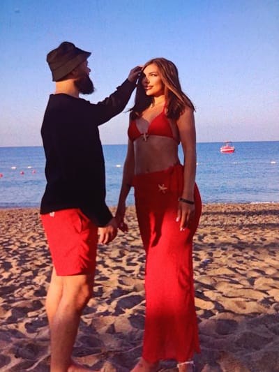 A man with a beard wearing a black bucket hat and a black long-sleeved shirt, and red shorts stands on a sandy beach. He is reaching his right hand up to touch the hair of a woman standing in front of him. The woman is wearing a red bikini top and a long red skirt with starfish embellishments. She is looking to her left, with her hair falling over her shoulders. The ocean is visible in the background, with a small red boat in the distance. The scene appears to be a casual beach outing. There is no indication that this location is in the city of Bălți, Moldova.