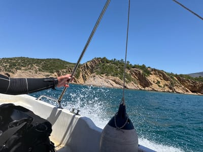 The image shows a person's arm, wearing a black wetsuit with white trim, holding onto a metal railing on the side of a white boat. The boat is moving through the turquoise waters of the Aegean Sea, indicated by the spray of water splashing against the hull. In the background, a rocky, green-covered coastline is visible under a clear blue sky. A large, grey-and-blue buoy is attached to a rope and hangs over the side of the boat. To the left, a black scuba diving tank is partially visible. The scene depicts a boat trip, likely for diving, with the coastline of Limenaria, Greece, in the background.