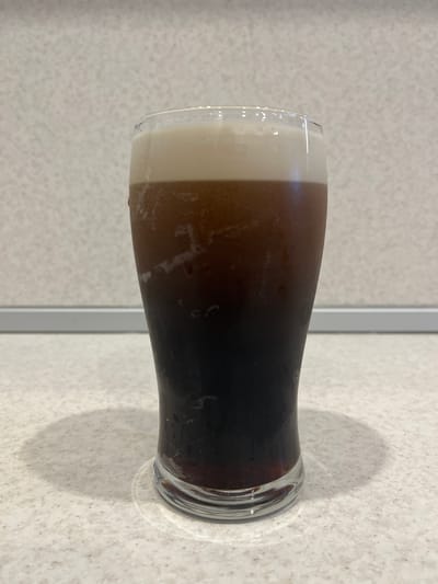 This media file is a photograph of a glass of dark beverage, possibly coffee or a stout beer, with a layer of lighter foam on top. The glass is tall and cylindrical, with a slight taper towards the base. The beverage itself is a deep, dark brown, almost black, and the foam is a creamy off-white. The glass is placed on a speckled, light-colored surface, which appears to be a countertop. A faint shadow of the glass is visible on the surface to the right. The background is out of focus, showing a wall with a textured pattern and a vertical element that might be a door frame or a partition. There are no people visible in the image. The lighting suggests an indoor setting, possibly during the daytime, given the natural light contributing to the shadow. The overall impression is one of a simple still life, focusing on the visual appeal of the drink. There is no visible text in the image. The location context of Azov, Russia, does not provide any specific visual cues within the image itself.