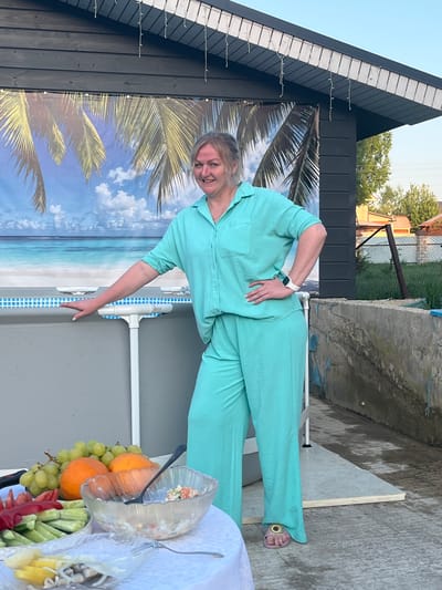 The image depicts a woman standing near what appears to be an above-ground pool, likely in a backyard setting. The location is identified as Новая Чернушка, Russia. The woman is dressed in a light blue pantsuit. Behind her is a backdrop featuring a beach scene with palm trees and a sky, attached to what looks like a shed or building with dark siding. A table with food, including fruit and salad, is in the foreground. The weather appears to be clear and sunny, based on the lighting.