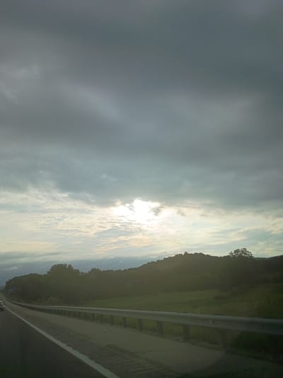 The image depicts a highway scene in Stanton, United States of America, under a cloudy sky with the sun partially visible. The perspective is from a moving vehicle, likely on the left side of the road, as indicated by the guardrail on the right. The highway stretches ahead, with a white line marking the lane. Beyond the guardrail, a green field slopes up to a tree-covered hill. The clouds are dark and textured, with patches of lighter sky and sunlight breaking through. The overall impression is of an overcast day with some illumination. There are no people or animals visible in the frame, and the focus is on the landscape and the road.