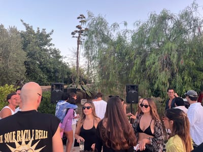 The image depicts an outdoor gathering at dusk, possibly at a private residence or venue in Fasano, Italy. A group of young adults are socializing, some holding drinks. Two black speakers are visible, suggesting music is playing. The background features lush greenery, including trees and a large agave plant. In the foreground, a man wearing a black t-shirt with a sun graphic and text is facing away from the camera. The overall atmosphere appears casual and festive.