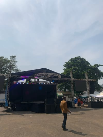 The image, oriented correctly, displays an outdoor event setup under a clear, bright daytime sky, likely in Freetown, Sierra Leone. A large, complex stage structure dominates the scene, constructed from metal trussing and covered in black fabric. Numerous speaker stacks are positioned on and around the stage, and a variety of colorful lighting fixtures, including LED par cans and moving heads, are mounted on the trussing, emitting blue, pink, and purple lights. A large, dark screen is visible on the upper left side of the stage structure.

In the immediate foreground, a man wearing a mustard yellow long-sleeved shirt and dark trousers stands with his back to the viewer, facing the stage. He holds a clear plastic bottle in his left hand, and his shadow is cast to his right on the paved ground. Lush green trees are visible behind and to the left of the stage, and another white tent structure, along with a few indistinct figures, can be seen in the background. A small white truck is partially visible near the lower left of the stage. A small, blurry green and white banner with unreadable text is in the far background.

The scene suggests the setup or a quiet moment before a public event, such as a concert, festival, or gathering, with the stage, sound system, and lighting prepared for use.