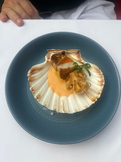 This image depicts a close-up, top-down view of a plated appetizer in a restaurant setting in Liverpool, United Kingdom. The central subject is a dish served within a natural, ridged scallop shell, which rests on a dark teal blue ceramic plate with a fine, textured, grid-like pattern. The scallop shell is predominantly white with brown striations along its outer edge.
Inside the shell, a dollop of smooth, vibrant orange-colored sauce or puree serves as a base. Positioned on this sauce is at least one seared scallop, exhibiting a golden-brown crust. A small cluster of bright orange-red roe (caviar) is placed adjacent to the scallop. To the right of the scallop and caviar, a light brown, crinkled, and ruffled edible garnish, possibly a fried mushroom or vegetable crisp, is present. A small sprig of green herbs with delicate leaves is placed atop this crinkled garnish. A hint of yellow material is visible beneath the scallop. At the bottom interior of the scallop shell, a few white granular crystals, consistent with coarse salt, are scattered.
The blue plate rests on a clean, untextured white tablecloth, which covers the majority of the visible surface. In the upper left quadrant of the image, the right hand of an individual is partially visible, resting flat on the white tablecloth. The hand exhibits visible knuckles and short, natural fingernails. Behind the hand, a portion of dark fabric, consistent with clothing, is discernible. The overall presentation suggests a formal dining experience.