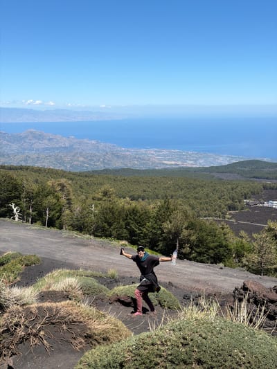 A person with a dark t-shirt, burgundy pants, and pink shoes stands on a dirt path on a mountainside, holding a plastic water bottle and a smartphone. The person is looking toward the camera with an open mouth. In the background, a vast green forest stretches out towards a hazy coastline and the blue ocean under a clear sky. The terrain is dark and rocky, with sparse vegetation in the foreground.
