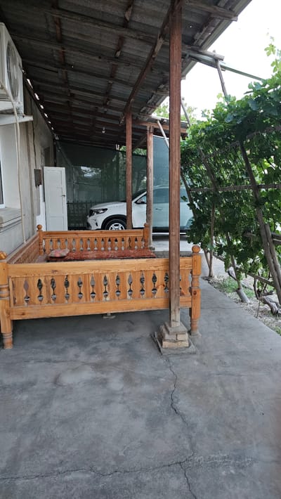 This media file depicts an outdoor scene in Gurlan, Uzbekistan, likely during the daytime with bright, overcast conditions. The primary focus is on a wooden swing, intricately carved with decorative patterns, positioned on a cracked concrete surface. The swing is placed beneath a rustic, open-sided structure with a corrugated metal roof supported by wooden beams. Lush green foliage, possibly grapevines, hangs from a trellis above the structure, suggesting a somewhat overgrown and natural setting.

In the background, a white car is partially visible, parked under the shelter. Further back, a white door or cabinet is visible. An air conditioning unit is also present in the lower left corner of the frame. The overall impression is one of a lived-in, possibly rural or semi-rural, outdoor space. The worn concrete and the utilitarian nature of the shelter suggest a practical use rather than an ornamental garden. The presence of the swing implies a recreational or relaxation area. There is no visible text in the image.