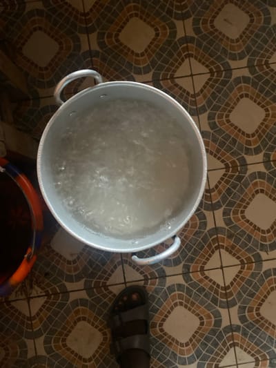 An overhead shot captures a domestic scene in Lakka, Sierra Leone, centered on a large, silver-colored metal pot, likely aluminum, containing actively boiling or bubbling water. The water's surface is frothy with numerous bubbles, indicating high temperature. The pot features two sturdy handles on opposite sides.

The pot rests on a tiled floor characterized by a repeating pattern of dark brown, beige, and light brown geometric shapes, creating a woven or basket-like appearance within square tiles. In the upper right quadrant of the frame, the lower part of a person's leg and foot are visible, clad in a dark-colored, two-strap sandal. The skin tone is dark. Partially visible in the bottom right corner is a section of a plastic bucket, displaying orange and possibly dark blue or black colors.

The primary activity depicted is the boiling of water, possibly for cooking, sanitation, or other household uses. The setting appears to be an indoor or shaded outdoor area, given the consistent lighting. No text is visible in the image.