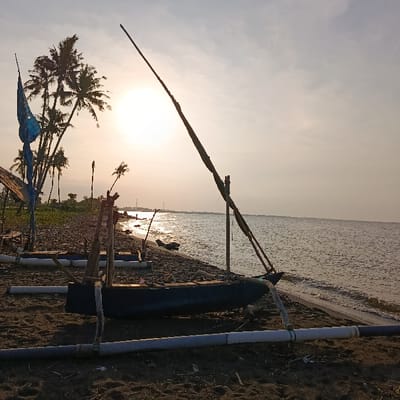The image shows a small wooden boat beached on a sandy shore in Arjasa, Indonesia, at coordinates -7.71 latitude and 114.118 longitude.  The boat is resting on its side with a long wooden pole extending upwards from its bow.  The pole appears to be a support for a sail, which is not visible in the image.  The boat is surrounded by other debris, including pieces of wood, rope, and plastic.  The sun is setting in the distance, casting a golden glow over the water. The shoreline is sandy and littered with debris.  The water is calm and clear, reflecting the colors of the sky. There are palm trees in the background and a few pieces of blue cloth in the foreground.  The sun is setting in the distance, casting a golden glow over the water.  A large wooden pole, perhaps a support for a sail, extends upwards from the bow of the boat.