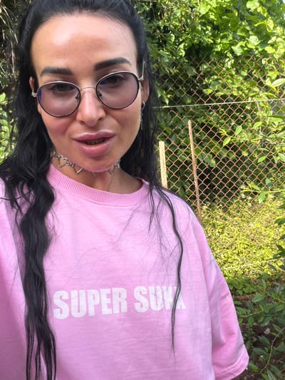 A medium shot captures a woman with long, dark hair, wearing round, wire-rimmed glasses and a pink sweatshirt with white lettering that reads "SUPER SUK." She has on a silver necklace with heart-shaped charms. The woman is looking towards the camera, with her mouth slightly open. Behind her, a chain-link fence covered in green foliage is visible. The setting appears to be an outdoor environment with lush vegetation, suggesting a park or garden in the city of Bronnitsy, Russia.