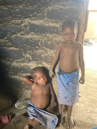 Two partially clothed children are lying on a dusty surface. The child on top is facing the camera with a neutral expression, while the other child is looking towards the camera with their arm resting on their head. Both children appear to be covered in dust or dirt. The surrounding environment seems to be a simple, possibly rural setting. The location is identified as Boi B, Nigeria.