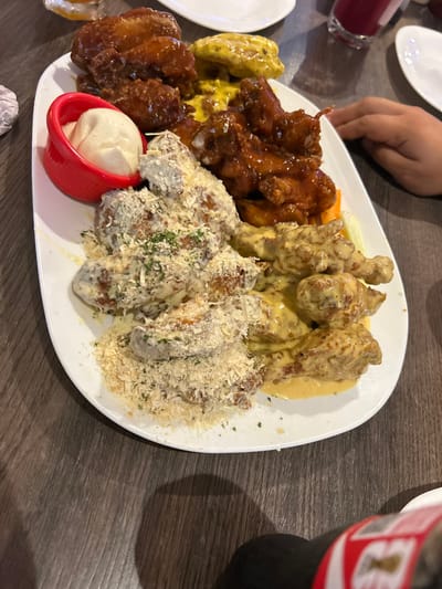 A platter of diverse chicken wings sits on a wooden table. The wings are coated in varying sauces and toppings, including a dark brown, yellow, creamy white with shredded cheese, and a mustard-like sauce. A small red container holds a white dipping sauce. A hand is partially visible, holding the edge of the plate. A bottle of Coca-Cola is partially visible on the right side of the frame. Other dishes and a glass are present in the background.