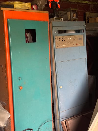 The image depicts a cluttered indoor space, possibly a workshop or storage area. Two large, retro-looking electronic cabinets dominate the foreground. The cabinet on the left is orange and teal, with a square opening revealing wires and what appears to be labeling inside. The cabinet on the right is blue, with a control panel featuring numerous buttons, dials, and a small screen. Other objects visible include what looks like a surveying instrument on top of the blue cabinet, a yellow strap, and some wooden planks in the background. The lighting suggests it is daytime, but the environment appears somewhat dim. There are no people or animals in the image. The location of Naro-Fominsk, Russia, is provided as context, but the image itself does not contain any visual cues to confirm this specific location.