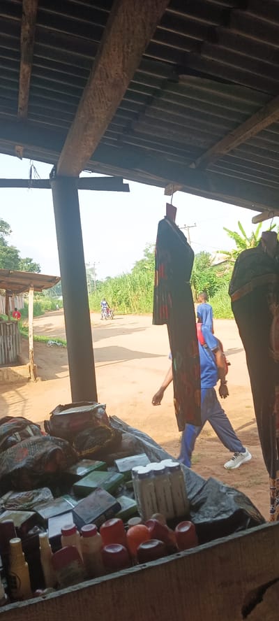 The image, taken from what appears to be a roadside shop in Amauzu, Nigeria, shows a dusty, dirt road with a person on a motorcycle in the distance. Closer to the camera, a young person wearing a blue shirt, grey pants, and white sneakers is walking along the side of the road. The foreground features a wooden shelf stocked with items wrapped in patterned fabric, along with bottles and boxes of goods, potentially for sale in the shop. The shop is covered by a corrugated metal roof supported by wooden beams. A couple of patterned fabric clothes hangs from the ceiling. Tall green vegetation lines the side of the road.