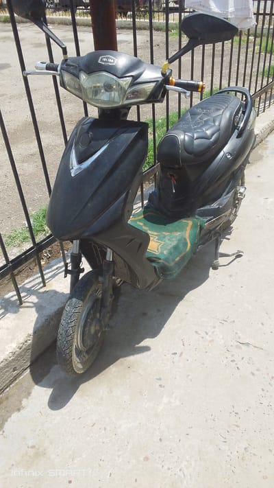 An eye-level, medium shot shows a black motor scooter parked on a concrete surface near a black metal fence in Dustabad (Soldatsky), Uzbekistan. The scooter is angled towards the viewer's left. The front of the scooter features a large headlight assembly with integrated turn signals. The handlebars are visible, with one grip covered in yellow tape and the other having a metallic end cap. The seat is black with a quilted pattern resembling a spiderweb. A green and gold patterned cloth is placed on top of the seat. The front wheel and part of the rear wheel are visible. The scooter is in a paved area with patches of grass growing through cracks in the concrete and along the base of the fence. The background shows a dirt or gravel area behind the fence, with some indistinct structures and a light-colored piece of fabric hanging. Shadows are cast on the concrete surface from the scooter and the fence. The image is sharp and well-lit, indicating it was taken during the daytime.