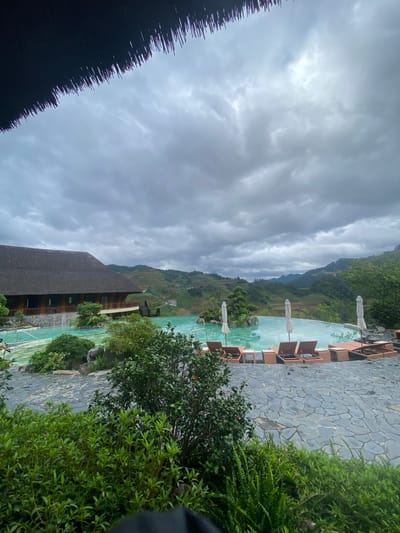 The image depicts an outdoor recreational area in Púng Luông, Vietnam, under an overcast sky. A large, irregularly shaped swimming pool with light turquoise water occupies the midground, featuring small islands with green foliage and rocks, including a sculpted miniature tree. Several brown lounge chairs with small side tables and white parasols (some open, some closed) are arranged around the pool's edge, which is paved with dark grey flagstones. To the left of the pool stands a multi-story building with a dark, possibly thatched roof and light-colored wooden walls, featuring balconies overlooking the pool and landscape. A wooden staircase is visible on the right side of the building. In the background, a series of green, terraced mountains and hills stretch towards the horizon under a heavily clouded, grey sky with some brighter patches. The foreground is partially obscured by the edge of a dark, thatched roof at the top and dense green bushes and foliage at the bottom. No distinct individuals are visible.