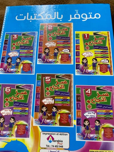 The image shows a blue textbook cover featuring a collage of six textbook covers in yellow, orange, red, purple, green, and brown. Each smaller cover is for "Success" and includes the semester, exercises, situations, choices, and a companion with precise corrections. Illustrations include children and subject names like "Mathematics," "Science," "French," and "Islamic Studies." The text on the blue cover says "Available in Libraries" in Arabic. A printing and publishing company logo, "Alarabia Print", is displayed with a telephone number and ISBN.