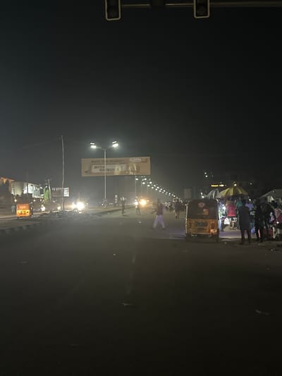 At night in Karum, Nigeria, a wide street is illuminated by overhead streetlights and the headlights of passing vehicles, casting long shadows and creating a somewhat hazy atmosphere. Pedestrians are visible on both sides of the road, some walking, others gathered in small groups. A yellow auto-rickshaw is parked on the right side of the street, and in the distance, several other vehicles are present. A large billboard displaying text and an image is suspended over the street on the left side, lit from below. The road surface appears to be asphalt, with some debris scattered along its edges. Buildings line the sides of the street, their facades dimly lit. The overall scene depicts a busy urban environment at night, with activity spilling onto the street.