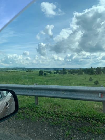The image captures a view from inside a vehicle, looking out over a vast, rolling landscape. The foreground is dominated by a metal guardrail, typical of a roadside, with green grass and gravel visible beneath it. To the left, the side-view mirror of the car reflects the brown exterior of the vehicle.

Beyond the guardrail, the scene opens up to a wide expanse of green fields dotted with scattered trees. In the distance, a dense line of trees forms the horizon, suggesting a forest or a wooded area. The sky above is a bright blue, filled with large, puffy white clouds, indicating a pleasant, possibly sunny day.

The overall impression is one of a rural or semi-rural setting, likely along a road in a natural environment. Given the prompt's mention of "Суда, Russia," this scene could be depicting the countryside surrounding the city, characterized by its rolling hills, green fields, and abundant trees under a dynamic sky. There are no people or specific structures clearly identifiable, suggesting a focus on the natural scenery and the experience of traveling through it.