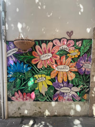 This image captures a vibrant mural painted on a textured, weathered wall in the city of Salerno, Italy. The mural depicts a profusion of colorful, stylized flowers in shades of pink, orange, yellow, blue, and purple, set against a black background. Many of the flowers have words written in white inside them, which appear to be Italian: "amicizia" (friendship), "amore" (love), "solidale" (solidarity), "pace" (peace), "rispetto" (respect), "salute" (health), and "insieme" (together). A heart shape is faintly visible on the wall above the mural, possibly a shadow or a separate drawing.

In the upper portion of the mural, a terra cotta planter with dry, dead foliage hangs directly above the flowers. To the left of the mural, a vertical metal pipe, likely a drainpipe or conduit, is attached to the wall, with several stickers visible on it. The wall itself shows signs of age and wear, with cracks and patches of discoloration, particularly at the bottom where it meets the sidewalk. The ground in front of the wall is paved with dark gray tiles, and scattered light and shadows, likely from overhead foliage, create a dappled effect on the wall and ground. The overall impression is one of urban art adding a splash of color and positive messages to a somewhat neglected corner of the city.
