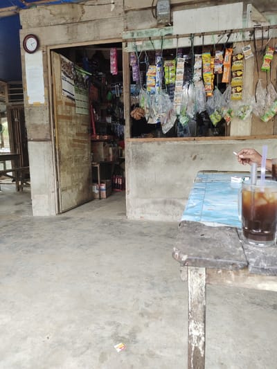 A weathered wooden storefront in Meunasahsagou, Indonesia, features shelves stocked with various snacks and beverages. In the foreground, a rustic wooden table holds a glass of iced coffee and a cigarette pack. A person's hand is visible, holding a lit cigarette. The ground is a concrete surface.