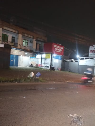 The image, captured at night in Ciomas, Indonesia, displays a street view featuring commercial establishments. The foreground shows an asphalt road with visible cracks and scattered debris. An elevated dirt and grass shoulder immediately borders the road, containing a white textile-like object and a blue plastic crate amidst sparse vegetation. A motorcycle in motion, characterized by significant blur and a visible red taillight, traverses the road from right to left.

To the right, a brightly illuminated red sign prominently displays "DOKTER 24 JAM WALI MEDIKA KLINIK PRATAPA," identifying a 24-hour medical clinic. Two dark motorcycles are parked on the unpaved ground directly in front of the clinic. A smaller yellow sign, partially obscured, reads "DIBUTUHKAN KONTRAKAN MELIHAT" (rental needed...viewing). Adjacent to the clinic on the left, a structure with blue corrugated roll-down doors exhibits a partially visible sign indicating "RUMAH MAKAN" (restaurant). One individual is seated on a bench or chair directly in front of this "RUMAH MAKAN" establishment. Above the structures, multiple utility lines are visible against the dark night sky. Artificial light sources illuminate the buildings and immediate surroundings.