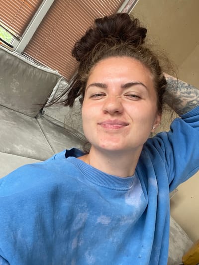 A young woman with dark hair pulled up in a messy bun is pictured in a close-up, frontal selfie. She is wearing a light blue crew neck sweatshirt with a tie-dye pattern. She has a light tan complexion with some redness on her cheeks and around her mouth. Her eyes are squinted, and she has a slight smile, with her right hand resting on the back of her head. Tattoos are visible on her right arm. The background shows a portion of a gray sofa, a window with brown blinds, and a wall. The scene appears to be indoors, possibly in a residential setting.