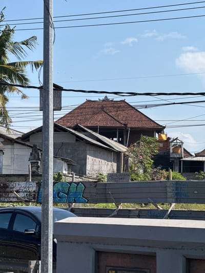 The image shows a street scene in Sukawati, Indonesia, on a bright, sunny day. The perspective is skewed, as if the camera is tilted. In the foreground, a metal railing runs horizontally across the frame, obscuring the lower portion of the scene. Behind it, glimpses of a dark blue vehicle and more metal structures are visible.
The mid-ground reveals a cluster of buildings with traditional tiled roofs, some appearing under construction or in disrepair. Wires crisscross the sky above, strung between poles. A lush green tree with pink flowers adds a touch of nature to the urban landscape. A sign with text is affixed to a vertical metal post.
Further back, more buildings are visible, along with vegetation. The sky is clear blue with a few wispy clouds, suggesting it is midday or early afternoon. The overall impression is of a busy, somewhat weathered, but still vibrant tropical locale. The visible text on the sign reads "JANGAN BUANG SAMPAH DI SINI," which translates to "Do not litter here" in Indonesian, indicating a public space. The graffiti on a metal surface adds a touch of urban art.