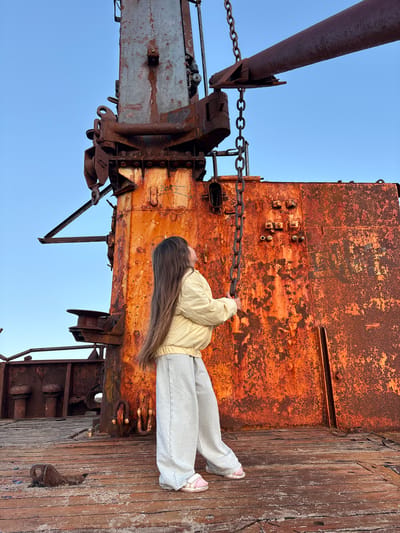 A young girl with long brown hair, wearing a light yellow jacket and grey pants, stands on the weathered wooden deck of a rusty ship. She is looking up at a thick metal chain hanging from a large, corroded industrial crane. The crane and the ship's structure are covered in a vibrant orange rust, contrasting with the clear blue sky. The scene appears to be set in the city of Teriberka, Russia, a location known for its ship graveyards and harsh coastal environment. The ground beneath her feet is also made of old wooden planks, showing signs of decay and wear.