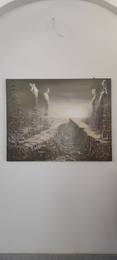The image shows a painting on a white wall, likely in a residential setting. The painting depicts a surreal and barren landscape with a deep chasm in the center. On either side of the chasm, there are fragmented structures resembling ruins and several ghostly, translucent figures. These figures appear to be busts or torsos, some with halos. The sky is overcast and somber. The painting is hung on a plain white wall, and a portion of the ceiling, which has a decorative molding, is visible at the top. A thin wire is protruding from the wall above the painting. The bottom of the image shows a speckled gray baseboard. The painting's surreal style suggests it might be influenced by artists like Salvador Dalí or Hieronymus Bosch. There is no indication of the location within the city of Voskevaz, Armenia.