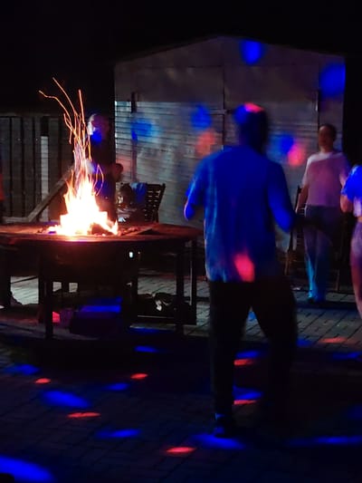 The image depicts an outdoor gathering at night, possibly in a rural area, given the mention of Мётчанскі сельскі Савет, Belarus, though the specific location cannot be definitively confirmed from the image alone. A bonfire is prominently featured in the foreground, burning brightly on a wooden table. Several individuals are present, their forms partially obscured by darkness and the dynamic lighting. One person in the mid-ground, facing away from the camera, wears a blue shirt and dark pants. To their right, another individual in a white shirt and dark pants is visible. Red and blue lights, likely from a disco ball or other lighting effects, are projected onto the ground and the surrounding structures, which appear to be a metal-clad building in the background. The ground is paved with interlocking bricks. The overall scene suggests a social event with music and dancing around a bonfire.