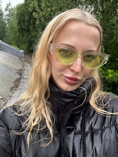 A young woman with blonde hair and yellow-tinted sunglasses poses for a selfie outdoors. She is wearing a black puffer jacket. The sunglasses have water droplets on them, suggesting it might be raining or has recently rained. Her blonde hair is long and slightly damp, with some strands falling over her face and onto her jacket. The background consists of lush green trees and a concrete path, indicative of an outdoor setting. The lighting appears to be natural daylight, possibly overcast due to the rain. The woman's expression is neutral, and she is looking towards the camera. There is no visible text in the image. The setting, with its greenery and outdoor elements, combined with the casual pose and attire, suggests a relaxed, everyday moment captured outside. The water droplets on the sunglasses are a prominent detail that hints at the weather conditions.