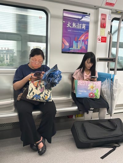 The image depicts two individuals seated on a train in Shuangfu, China. The train's interior features metal seating, windows with adjustable blinds, and advertisements. On the left, a woman with glasses and dark hair is engrossed in her smartphone. She is wearing a dark blue t-shirt and black pants, and her feet are clad in black Crocs with decorative charms. Beside her, she has a large tote bag adorned with a floral pattern and cartoon characters, and a blue umbrella. To the right, a young woman with long dark hair is also looking at her smartphone. She is wearing a light pink collared shirt and a dark skirt with white socks. In front of her is a gift bag with the words "BEST WISH THANKS YOU LOVE" and floral decorations, along with a clear umbrella. On the floor in front of the young woman rests a black duffel bag. The overall scene suggests a quiet commute.
