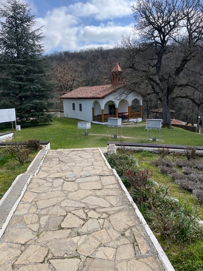 The image depicts the courtyard of a religious complex located in the city of Zlatna Livada, Bulgaria. A stone pathway constructed from irregular flagstones, bordered by concrete, extends from the foreground towards a small, white-walled building with a terracotta-tiled gable roof and an ornate copper-colored bell tower. The building features an arched portico with wooden columns and a railing, revealing dark double doors, a table with a red cloth, and benches inside.
The pathway is flanked by garden beds containing dormant shrubs and green plants. Beyond the path, a manicured green lawn occupies the midground. On the lawn, in front of the building, three grey, rectangular metal stands with cross-shaped cutouts on top are visible, each positioned over a small bucket. To the left of the building, a tall evergreen conifer stands beside a white rectangular informational sign on a stand. To the right of the building, a large deciduous tree with bare branches dominates the space. Additional low shrubs and two dark-colored benches are partially visible on the right side of the lawn. The background consists of a dense forest of deciduous trees covering a hillside under a partly cloudy blue sky. No individuals are present.