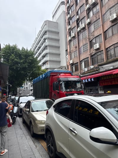 A street scene in Liwan District, China, shows a heavily overcast sky over multi-story buildings lining a narrow street. In the foreground, parked vehicles, including a red truck with a blue tarp covering its cargo, a silver sedan, and a white SUV, are visible. A person, carrying a child, walks on the sidewalk. The buildings feature numerous windows and air conditioning units. Shops with red awnings are visible on the right side of the street. The overall impression is of a busy urban environment.