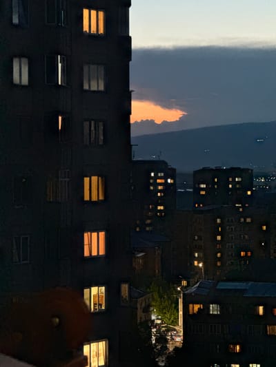 The image captures a city skyline at dusk, likely in Abovyan, Armenia, with a prominent apartment building in the foreground on the left. The building's dark facade is punctuated by numerous windows, some illuminated by warm, golden light, suggesting occupancy. A few windows are dark or dimly lit. The sky above is a gradient of fading daylight, with a band of dark clouds obscuring a portion of a vibrant orange sunset. In the background, several other apartment buildings are visible, also displaying a scattering of lit windows, indicating a residential urban environment. A street with car lights is visible in the middle ground, and distant city lights twinkle on the horizon, suggesting a larger urban sprawl beyond the immediate foreground. The overall atmosphere is one of a quiet evening settling over a densely populated area.