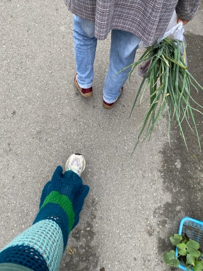 The image shows a low-angle shot of two people standing on a grey asphalt surface, likely a street or sidewalk in Chongzhou City, China. The person in the foreground is wearing a white sneaker with light purple accents and a blue, green, and teal crocheted leg warmer. They are holding a dark green object that appears to be part of their clothing. The person in the background is wearing blue jeans, red shoes with a brown sole, and a plaid jacket. They are holding a bunch of green onions in a clear plastic bag. To the right of the background person, a blue plastic basket containing green leaves is visible. The asphalt surface appears wet in some areas, suggesting recent rain.