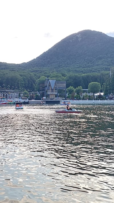 The image, rotated 90 degrees counter-clockwise, depicts a scenic waterscape with a backdrop of a lush, tree-covered mountain. In the foreground, the sun glints off the choppy water, creating a shimmering effect. A red and white paddleboat, carrying at least three individuals wearing life vests, is being propelled across the water. To the left of the water, a wooden pier extends into the lake, lined with small lights. Several small buildings and structures are visible along the shore, including a multi-story building with a distinctive angular roof and balconies. The overall atmosphere suggests a leisurely summer day, possibly late afternoon or early evening, given the quality of light. The location appears to be a recreational area, likely a lake or reservoir, in Zheleznovodsk, Russia, given the context. The presence of boats and the setting imply an outdoor activity such as boating or swimming. There is no visible text in the image.