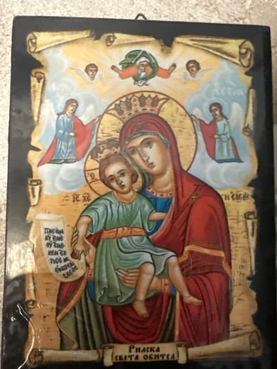 This is a photograph of an icon depicting the Virgin Mary holding the Christ child. They are surrounded by celestial figures and scrolls with writing. The icon is framed by a black border and has a dark, textured background. The style is traditional Orthodox Christian iconography. The photograph was taken in Marten, Bulgaria.