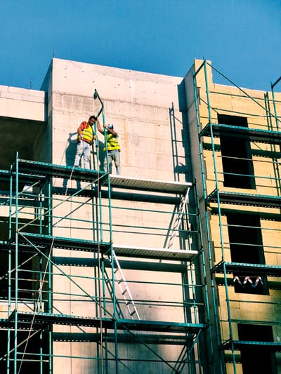 Two construction workers in high-visibility vests are working on the exterior of a building under construction. The building appears to be made of concrete and scaffolding, with wooden panels and metal supports. The scene is set against a clear blue sky, suggesting it is daytime. The workers are positioned at different heights, one above the other, and are secured by safety harnesses. They are using the scaffolding to access different parts of the building. One worker is leaning on a railing, while the other is holding onto a support beam. The sun is casting strong shadows on the building, indicating it is likely a sunny day. There are no visible signs of text in the image. The location is Đenovići, Montenegro.