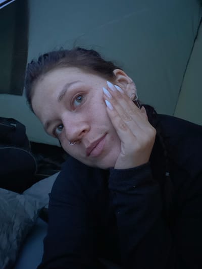 The image shows a close-up, self-taken photograph of a young woman's face, partially obscured by her hand. She has light skin with freckles, green eyes, and light pink lips. Her fingernails are long and painted white. She is wearing a dark-colored top and has a nose ring. In the background, a tent's interior is visible, with a sleeping bag or mat on the left. The overall lighting is dim, suggesting an indoor or low-light environment. The location is identified as Абзаново, Russia.
