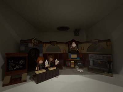 This image, originating from McAllen, United States of America, displays a LEGO diorama meticulously arranged within a plain, off-white enclosure, likely a shelf or display cabinet. The diorama itself is a dimly lit, brick-built room, suggesting a classroom or study environment.
In the foreground, two LEGO minifigures, both attired in dark robes and possessing brown hair, are positioned at individual brown desks. The minifigure on the left holds a light-colored implement, resembling a quill, poised over a tan flat tile that represents a book or scroll. The minifigure on the right holds a white, feather-like element over a similar tan tile. To the left of these figures, a brown LEGO blackboard, supported by a reddish-brown easel, displays white chalk markings depicting celestial bodies (a crescent moon and stars), swirls, a feather, and a pointed, wand-like object.
In the mid-ground, a third LEGO minifigure, an elderly male with a prominent white beard and brown robes, stands behind a brown podium or lectern. Behind this figure, a tiered bookshelf, constructed from brown LEGO bricks, houses various colorful LEGO brick 'books.' To the right, a gray LEGO fireplace with an ornate mantelpiece is prominent. The fireplace opening contains orange and red bricks simulating flickering flames, and an open book rests above the mantelpiece. A white, serpentine LEGO element lies on the floor directly in front of the fireplace. The background wall of the diorama incorporates arched structures and decorative elements, including crests or shields mounted above. The overhead interior of the display enclosure reveals a dark circular vent and a small, dark rectangular fixture.