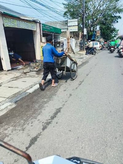 A man wearing a blue shirt and khaki pants is pushing a handcart down a paved street. The handcart is loaded with goods. The man is walking on the right side of the street, which is clear of other pedestrians or vehicles. The man is facing the street and looking ahead. There are buildings and trees on both sides of the street. The scene is captured in Cimahi, Indonesia, at latitude -6.894 and longitude 107.559. The street is paved, with some slight depressions in the surface. There is a sign above the man's head that reads "Cuci Motor 244." There are several other vehicles parked on the side of the street, including a car, a motorcycle, and a van. There are a few people standing or walking in the background. The day is sunny and the air is clear.
