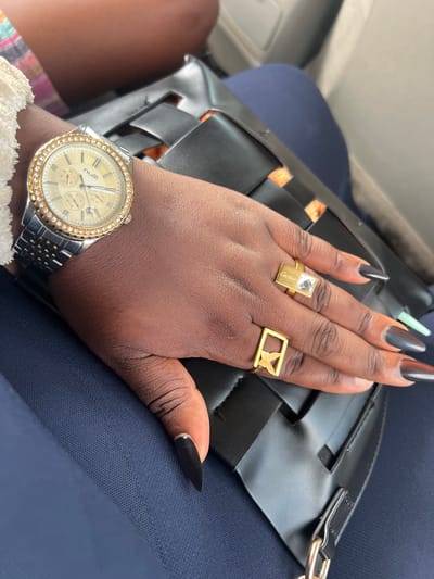 A person's left hand and wrist are prominently displayed in the image. The person has dark skin and wears a watch on the wrist with a silver band and a gold face surrounded by small gems. Two rings are visible on the hand: one is a rectangular gold ring with a clear gem, and the other is a rectangular gold ring with a butterfly design. The fingernails are long, pointed, and painted black. The hand is resting on a black woven purse or bag placed on the person's lap, which is covered with blue fabric. The location is not apparent in the image, and there are no details to suggest it is in Nabatum, Nigeria.