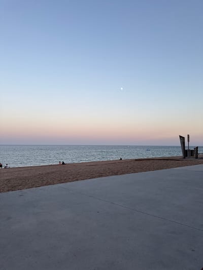 This image depicts a beach scene at Platja D'aro, Spain, during what appears to be dusk or dawn due to the soft, gradient lighting in the sky transitioning from pale blue to a warm peach hue. A bright, small moon is visible in the upper left portion of the sky. The scene is divided diagonally, with the left side dominated by the vast expanse of the sky and the ocean, and the right side by a textured, grey concrete promenade or walkway.

Along the edge of the water, a sandy beach is visible, with a few small figures scattered across it, suggesting people are present, though their activities are not clearly discernible. Some appear to be sitting or standing near the water's edge. In the water itself, there are small ripples, indicating a calm sea. Further out, a couple of small, blue objects are visible in the water, possibly buoys or small inflatable items.

Along the promenade, there are some metallic structures that look like part of a seating area or perhaps some kind of equipment. The concrete promenade has visible lines or seams, and its surface is a neutral grey. The overall impression is one of a tranquil evening or early morning at a coastal location, with the soft light creating a peaceful atmosphere. There is no visible text in the image.