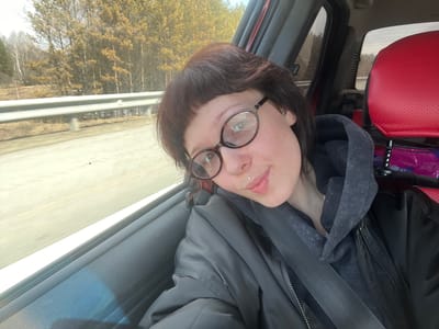 A woman wearing glasses and a black jacket is sitting in the passenger seat of a car. She has short, dark hair and a lip piercing. She is wearing a gray hoodie under the jacket. The seat next to her is red. Outside the car window is a highway with a guardrail and trees. The car is traveling in Russkie Kirmeni, Russia.