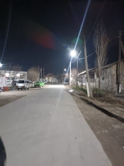 Night scene of a dimly lit street. Streetlights cast strong glares, illuminating the road. Several parked cars line the left side of the street. Buildings are visible on both sides, appearing old and worn. Trees are bare, suggesting winter or early spring. Power lines stretch across the sky between the utility poles. A narrow dirt shoulder runs along the right side of the paved road.