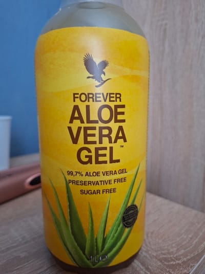 The scene depicts an indoor environment. No visual indicators are present to identify a precise location within Selyanin, Bulgaria, or any other specific geographical area.
The primary subject is a tall, cylindrical bottle of "FOREVER ALOE VERA GEL," positioned upright and slightly angled, occupying the majority of the frame. The bottle features a bright yellow label with prominent dark brown text. A stylized dark brown eagle logo is visible near the top of the label, above the word "FOREVER." Below this, "ALOE VERA GEL™" is displayed. Further down, the label states "99.7% ALOE VERA GEL," "PRESERVATIVE FREE," and "SUGAR FREE." A graphic depiction of green aloe vera leaves is present on the lower part of the label, extending towards the base, where "1L" is visible, indicating volume. The top of the bottle, above the label, appears to be clear or transparent plastic.
The bottle rests on a light-colored, possibly wooden, horizontal surface exhibiting a faint grain pattern. To the left of the bottle, in the mid-background, a partial view of a light blue surface is visible, along with a segment of a rose-gold metallic object, possibly an electronic device. The background to the right of the bottle consists of a vertical surface with a light wood grain pattern. No individuals are present in the image.
